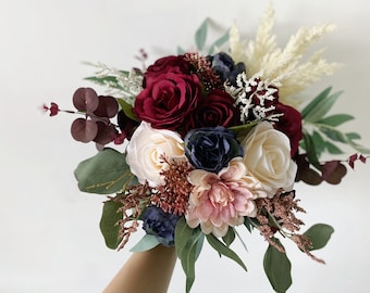 May include: A wedding bouquet with burgundy roses, ivory and navy blue blooms, and pampas grass. The arrangement includes eucalyptus leaves and other decorative elements, creating a romantic and elegant look.