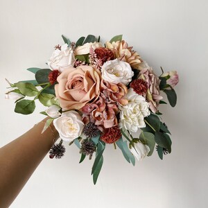 May include: A bouquet of artificial flowers in shades of blush, cream, and burgundy. The bouquet includes roses, dahlias, and eucalyptus.
