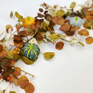May include: A garland of brown eucalyptus leaves with small white flowers and three miniature pumpkins, one orange and two green.