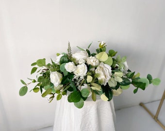 May include: A white floral arrangement featuring white roses and greenery. The arrangement is displayed on a white tablecloth.