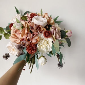 May include: A bouquet of artificial flowers in shades of peach, white, and burgundy. The bouquet includes roses, peonies, and other floral elements.