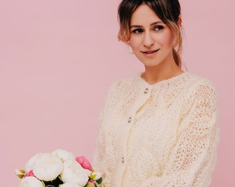 May include: A woman wearing a white knitted cardigan and white trousers holds a bouquet of white and pink peonies against a pink background.