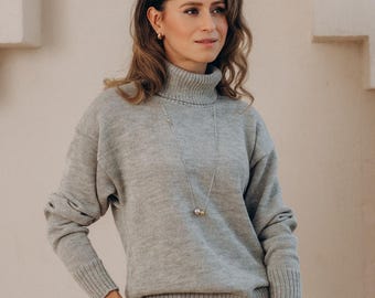 May include: A woman wearing a gray turtleneck sweater and white pants. She is also wearing a long silver necklace. The background is a light beige wall with an arched architectural detail.