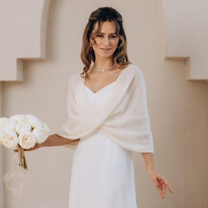 Puede incluir: Una novia con un vestido de novia blanco y un chal sostiene un ramo de flores blancas. El vestido es largo hasta el suelo con cuello en V. El chal es de color blanco roto claro. El fondo es una pared beige claro con un diseño arqueado.