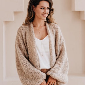 May include: A woman wearing a light brown knitted cardigan with long sleeves and a white tank top. She is wearing brown pants and has long brown hair. She is standing in front of a white wall with an arched doorway.