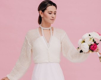 Può includere: Una sposa indossa un cardigan bianco di maglia sopra un abito bianco, tenendo un bouquet di fiori bianchi e rosa. È in piedi davanti a uno sfondo rosa.