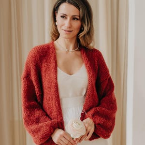 May include: A rust-colored, open-front cardigan with a chunky knit texture. The cardigan is worn over a white dress. The image features a woman holding a light pink flower. The background is a neutral beige.