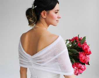 May include: A white knitted bridal shawl with a low back and a gathered detail in the back. The shawl is worn by a woman with dark hair and a bouquet of pink peonies.