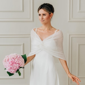 Puede incluir: Una novia con un vestido de novia blanco y un chal blanco transparente, sosteniendo un ramo de hortensias rosas. El vestido tiene un corpiño ajustado y una falda fluida. El chal añade un toque de textura delicada.