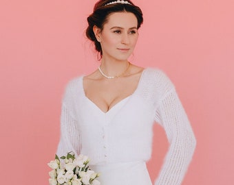 Può includere: Una donna con un cardigan di maglia bianca e una gonna tiene un mazzo di fiori bianchi. Indossa una collana di perle e un cerchietto di perle. Lo sfondo è rosa solido.