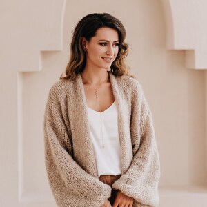 May include: A woman wearing a light brown knitted cardigan with long sleeves and a white top. She is standing in front of a white wall with an arched doorway.
