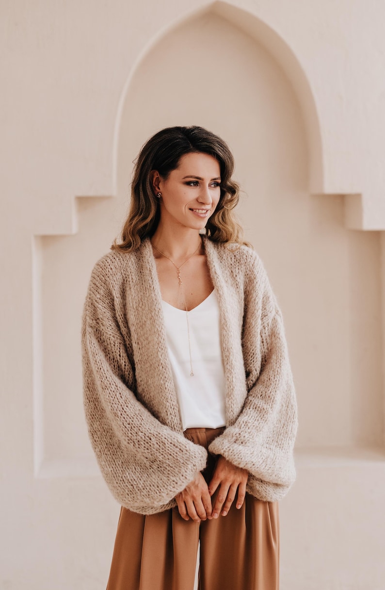 May include: A woman wearing a light brown knitted cardigan with long sleeves and a white top. She is standing in front of a white wall with an arched doorway.