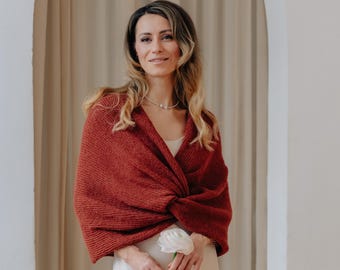 May include: A rust-colored knitted wrap featuring a textured stitch pattern and a twisted front design. The garment is worn over a simple cream-colored slip dress, providing a layer of warmth with its soft, woven fabric construction.