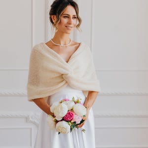 May include: A cream-colored knitted shrug featuring a soft, fuzzy texture and a central twisted knot detail. The garment is worn over a plain white dress, with the knit fabric draped across the shoulders and upper arms, creating a layered look.