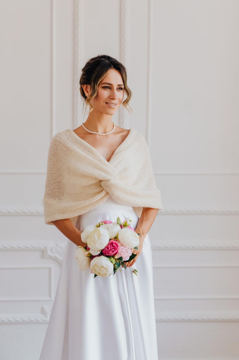 May include: A cream-colored knitted shrug featuring a soft, fuzzy texture and a central twisted knot detail. The garment is worn over a plain white dress, with the knit fabric draped across the shoulders and upper arms, creating a layered look.