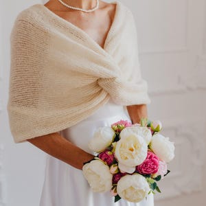 May include: A cream-colored, knitted shrug featuring a front twist detail and short sleeves, worn over a plain white dress. The garment is paired with a pearl necklace and a bouquet of white and magenta peonies with green foliage.