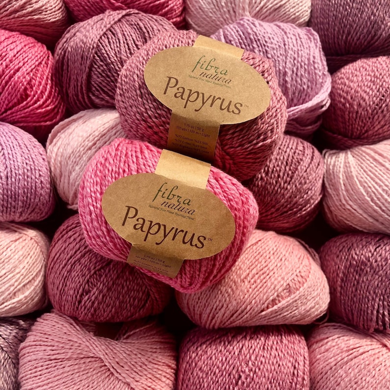 May include: A close-up of a pile of pink yarn balls with brown labels that read "fibra natura Papyrus".