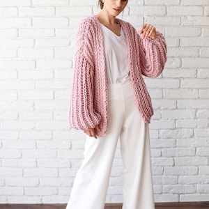 Pu&ograve; includere: Una donna indossa un cardigan di maglia rosa con maniche ampie e pantaloni bianchi larghi. &Egrave; in piedi davanti a un muro di mattoni bianchi.