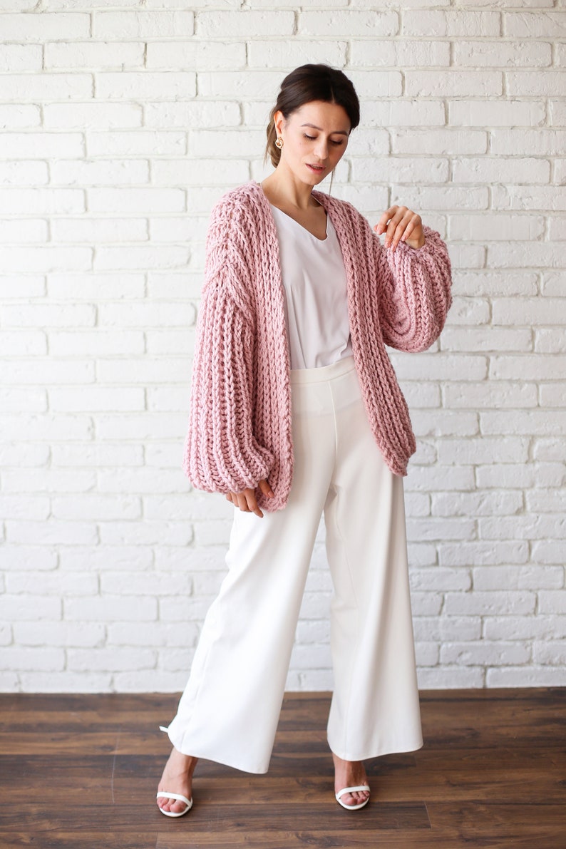 Pu&ograve; includere: Una donna indossa un cardigan di maglia rosa con maniche ampie e pantaloni bianchi larghi. &Egrave; in piedi davanti a un muro di mattoni bianchi.