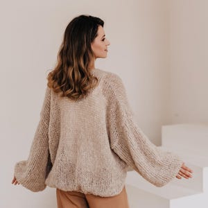 May include: A woman wearing a light brown, oversized knitted cardigan with a loose fit and long sleeves. She is wearing brown pants and standing in front of a white background.