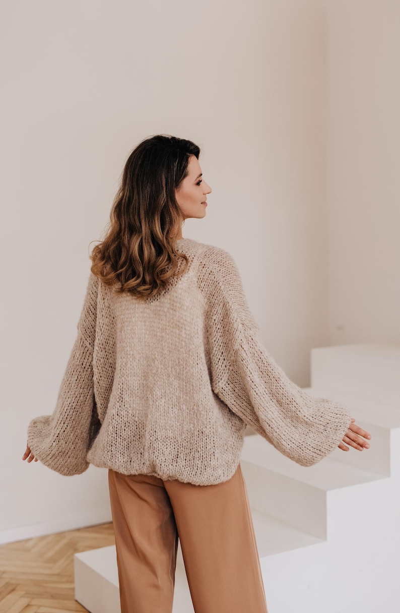 May include: A woman wearing a light brown, oversized knitted cardigan with a loose fit and long sleeves. She is wearing brown pants and standing in front of a white background.