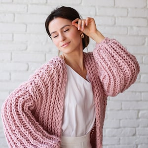 Pu&ograve; includere: Una donna indossa un cardigan di maglia rosa con maniche larghe a maglia grossa. Il cardigan &egrave; aperto e la donna indossa una camicetta bianca e pantaloni bianchi.