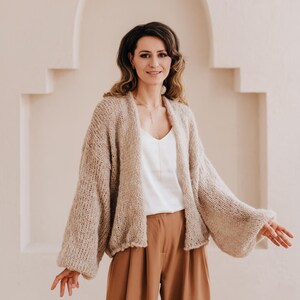 May include: A woman wearing a light brown knitted cardigan with a white top and brown wide-leg pants. She is standing in front of a white wall with an arched doorway.