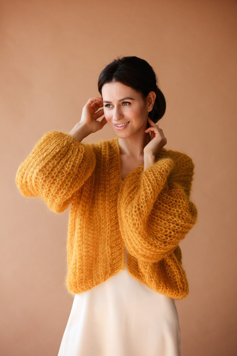 May include: A woman wearing a yellow knitted cardigan with large sleeves. The cardigan is made of a soft, fluffy yarn and has a relaxed fit.