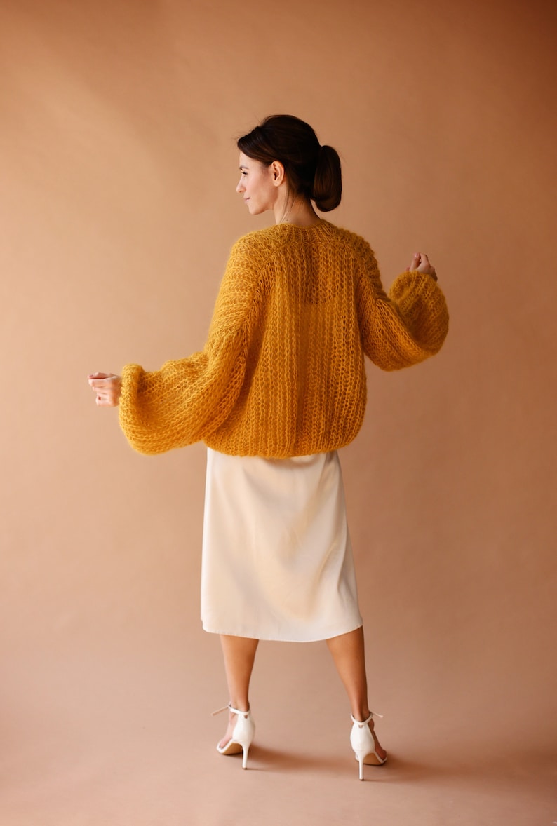 May include: A woman wearing a mustard yellow knitted cardigan with a white skirt and white heels.