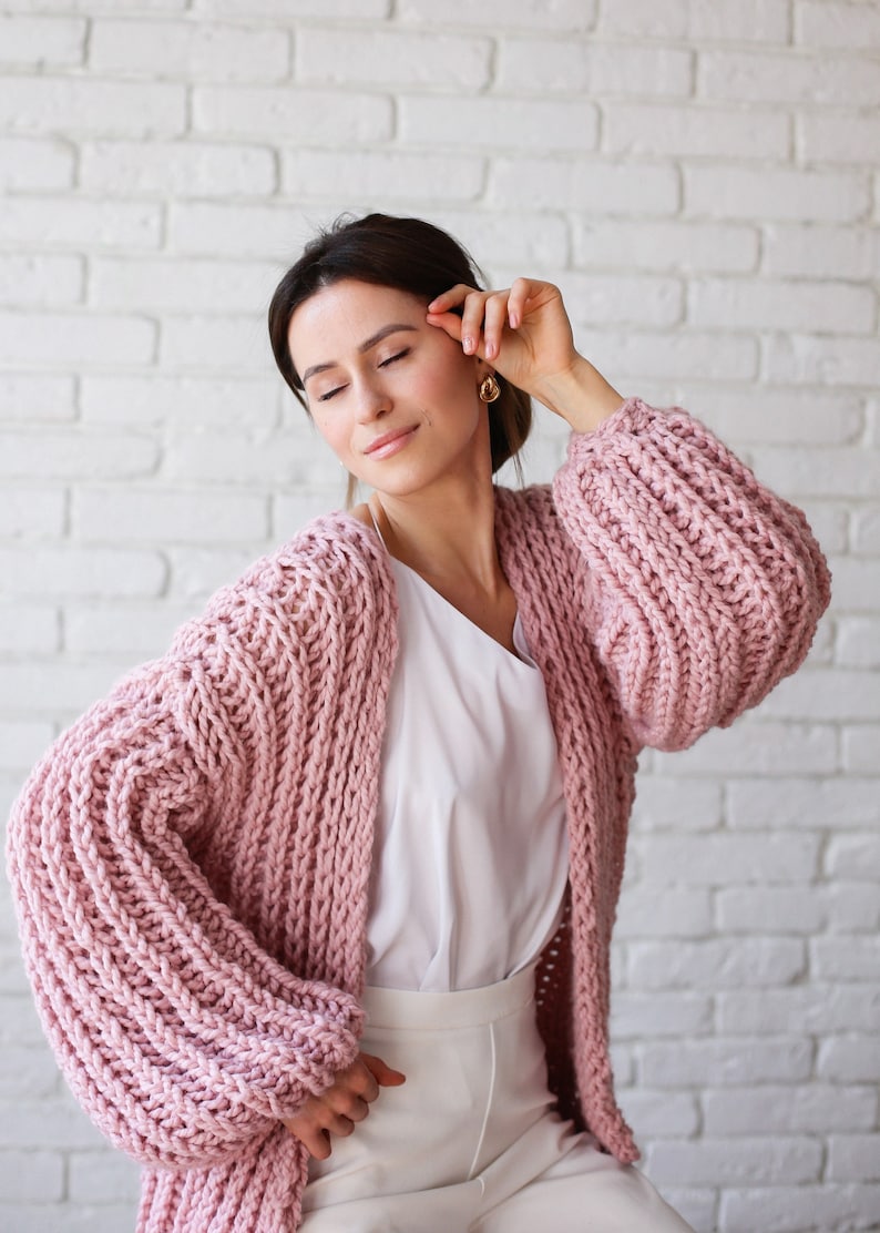 Pu&ograve; includere: Una donna indossa un cardigan di maglia rosa con maniche larghe a maglia grossa. Il cardigan &egrave; aperto e la donna indossa una camicetta bianca e pantaloni bianchi.