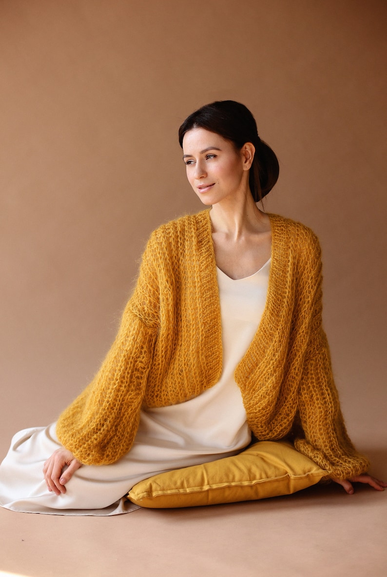 May include: A woman wearing a yellow knitted cardigan with long sleeves and a white slip dress. She is sitting on a yellow cushion with her legs crossed.