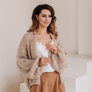 May include: A woman wearing a light brown knitted cardigan with a white tank top and brown wide-leg pants.