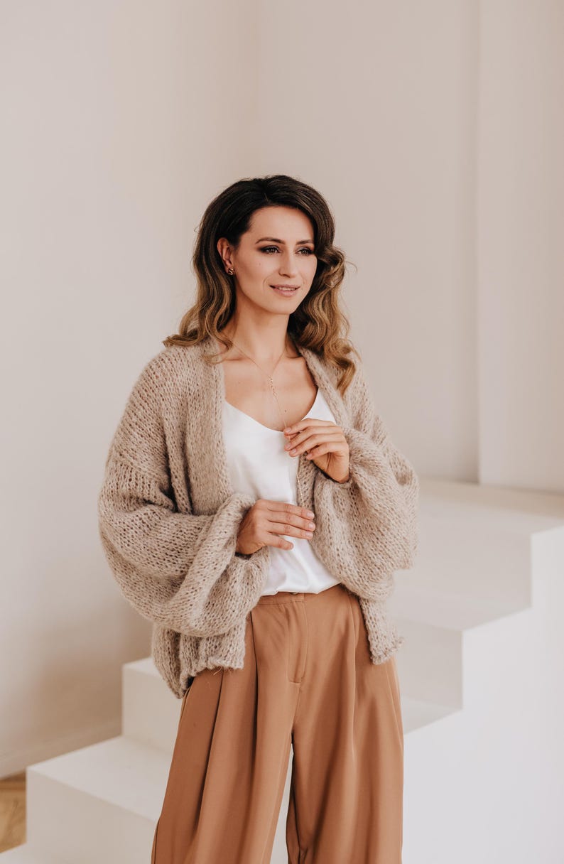 May include: A woman wearing a light brown knitted cardigan with a white tank top and brown wide-leg pants.