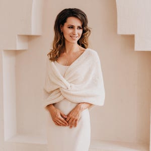 May include: A cream-colored, knitted shrug featuring a twisted front design and long sleeves. The garment is made of a soft, textured yarn and is worn over a simple, sleeveless, floor-length dress in a matching light cream shade.