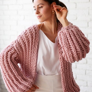Pu&ograve; includere: Un cardigan di maglia rosa con maniche grandi e gonfie. Il cardigan &egrave; aperto davanti e viene indossato sopra una canottiera bianca e pantaloni bianchi.
