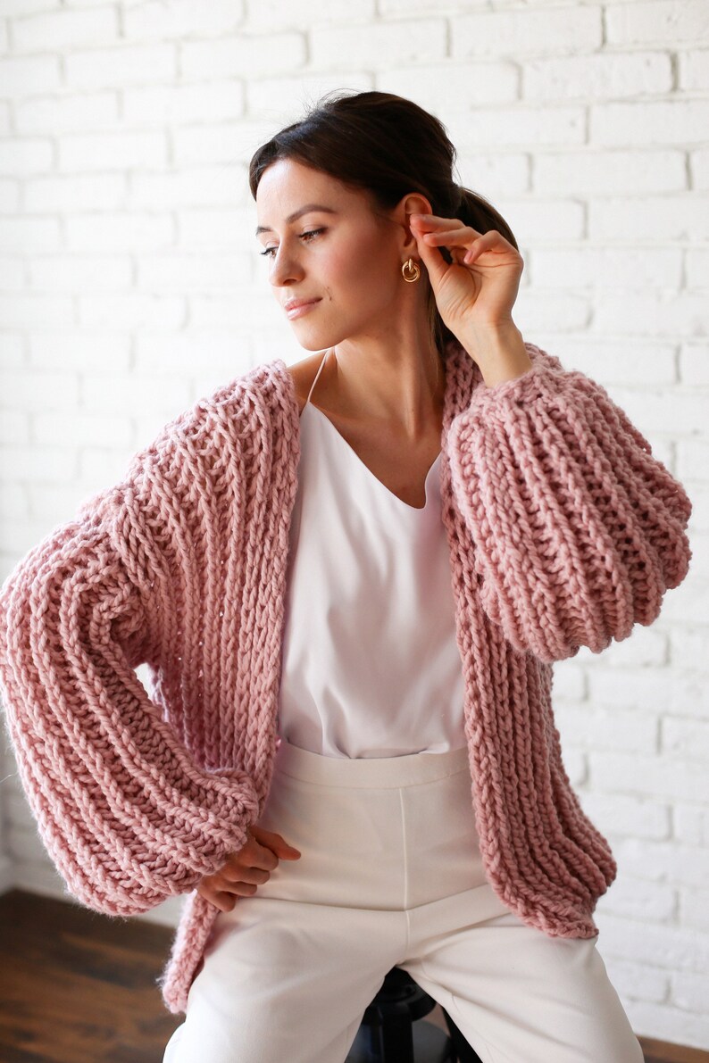 Pu&ograve; includere: Un cardigan di maglia rosa con maniche grandi e gonfie. Il cardigan &egrave; aperto davanti e viene indossato sopra una canottiera bianca e pantaloni bianchi.