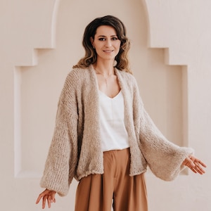 May include: A woman wearing a light brown knitted cardigan with a white top and brown wide-leg pants. She is standing in front of a white wall with an arched doorway.