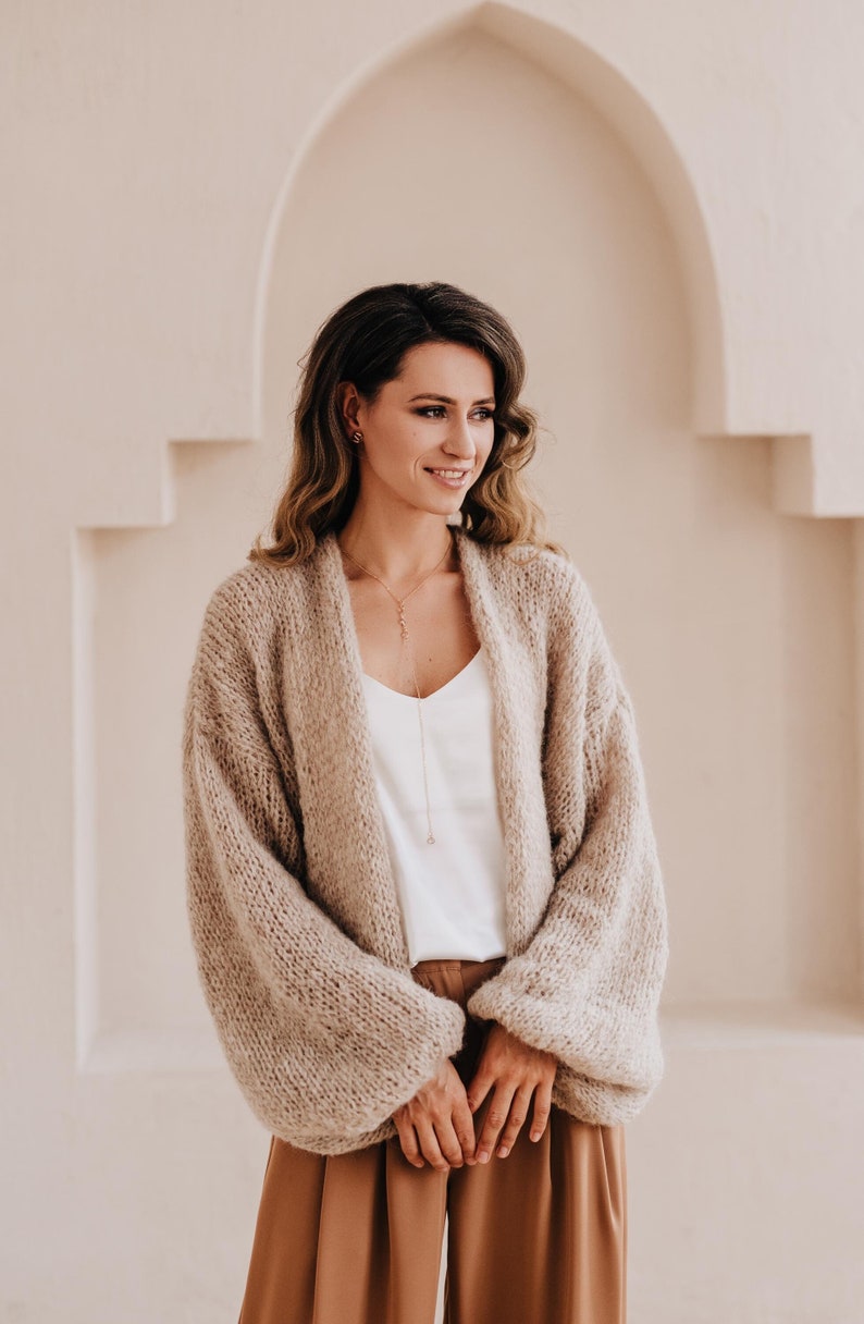May include: A woman wearing a light brown knitted cardigan with long sleeves and a white tank top. She is wearing brown pants and has long brown hair. She is standing in front of a white wall with an arched doorway.