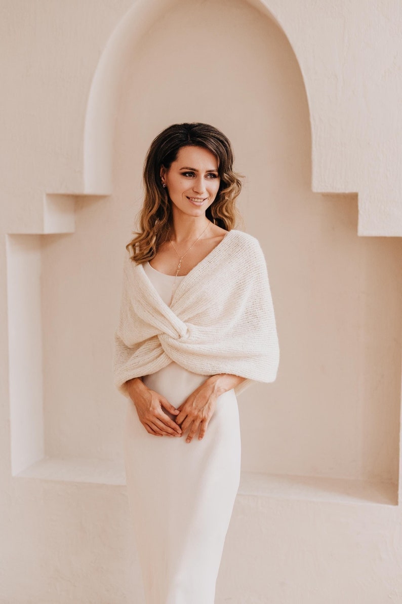 May include: A cream-colored knitted shrug featuring a twisted front design and long sleeves. The garment is made of a soft, textured yarn and is worn over a matching cream-colored sleeveless dress, creating a cohesive and layered appearance.