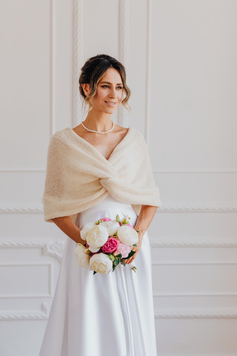 May include: A cream-colored knitted shrug with a twisted front design worn over a white dress. The garment features a soft, textured knit fabric with short sleeves and a V-neckline. A bouquet of white and pink peonies is held in front of the dress.