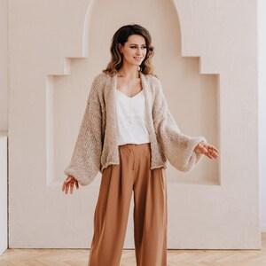 May include: A woman wearing a beige knitted cardigan with large sleeves and brown wide-leg pants. She is standing in front of a white wall with an arched doorway.