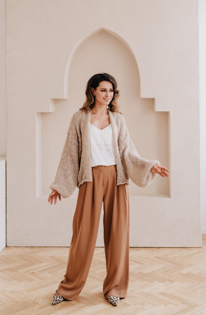 May include: A woman wearing a beige knitted cardigan with large sleeves and brown wide-leg pants. She is standing in front of a white wall with an arched doorway.