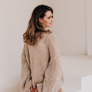 May include: A woman wearing a light brown, chunky knit sweater with long sleeves and a relaxed fit. She is wearing brown pants and is standing in front of a white wall.