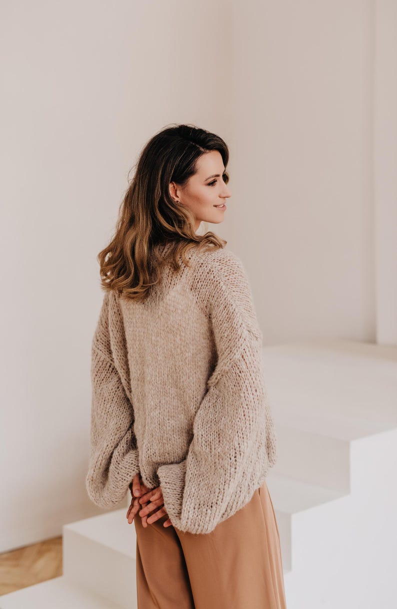 May include: A woman wearing a light brown, chunky knit sweater with long sleeves and a relaxed fit. She is wearing brown pants and is standing in front of a white wall.