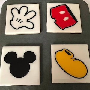 May include: Set of four ceramic coasters featuring iconic Mickey Mouse imagery. The coasters depict Mickey's glove, shorts, head, and shoe in black, white, red, and yellow.