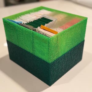 Könnte beinhalten: Ein zweifarbig grüner Teebeutel-Organizer mit einem strukturierten Blättermuster. Der obere Teil ist hellgrün, während der untere Teil einen dunkleren Farbton aufweist. Die Box ist mit verschiedenen Teebeuteln gefüllt, darunter einige mit der Marke "TAZO".