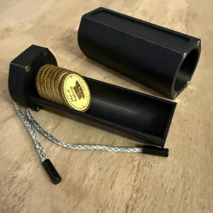 May include: Black hexagonal coin holder with a matching lid. The open holder reveals several gold coins with the text "Hat of Sighing". A silver and white cord is attached to the holder.
