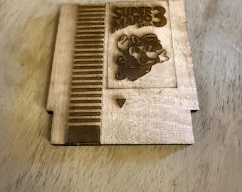 Könnte beinhalten: Holzuntersetzer in Form einer klassischen Videospielkassette. Der Untersetzer zeigt den Text "SUPER MARIO 3 BROS." und ein Bild von Mario. Der Untersetzer ist aus hellem Holz gefertigt.