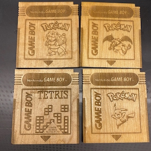 May include: Four wooden Game Boy cartridge coasters featuring the classic Nintendo Game Boy logo and various game titles, including Pokemon, Tetris, and Pokemon. The coasters are made of light brown wood and have a natural wood finish.