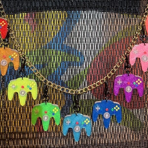 May include: A gold chain necklace with several small, colourful game controller pendants. The controllers are in various shades, including red, orange, green, blue, and purple, with detailed button designs. The background is a mesh fabric with a colourful graffiti design.